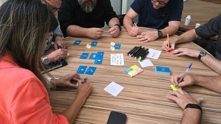 Equipe diversa reunida em torno de uma mesa, discutindo ideias e organizando cartões e notas adesivas durante uma sessão de brainstorming.